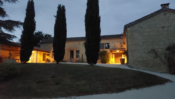 A stone building with a courtyard, tall cypress trees, and a warm-lit entrance.