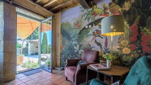 A room with a large mural of birds and flowers, a leather armchair, a table with a lamp, and a view through a glass door to an outdoor area with a stone wall and plants. - Duplex studio "La Volière" 10 minutes from Lectoure, Fleurance and Condom (Terraube)