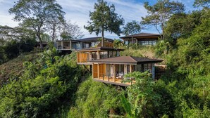 Ein modernes, mehrgeschossiges Haus mit großen Glasfenstern, Holzterrassen und üppiger, umgebender Bepflanzung.