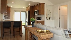 A modern kitchen with wooden cabinets, a stainless steel refrigerator, and a built-in microwave. There is a wooden bar counter with two stools and a small potted plant. A light-colored sofa is visible in the background.