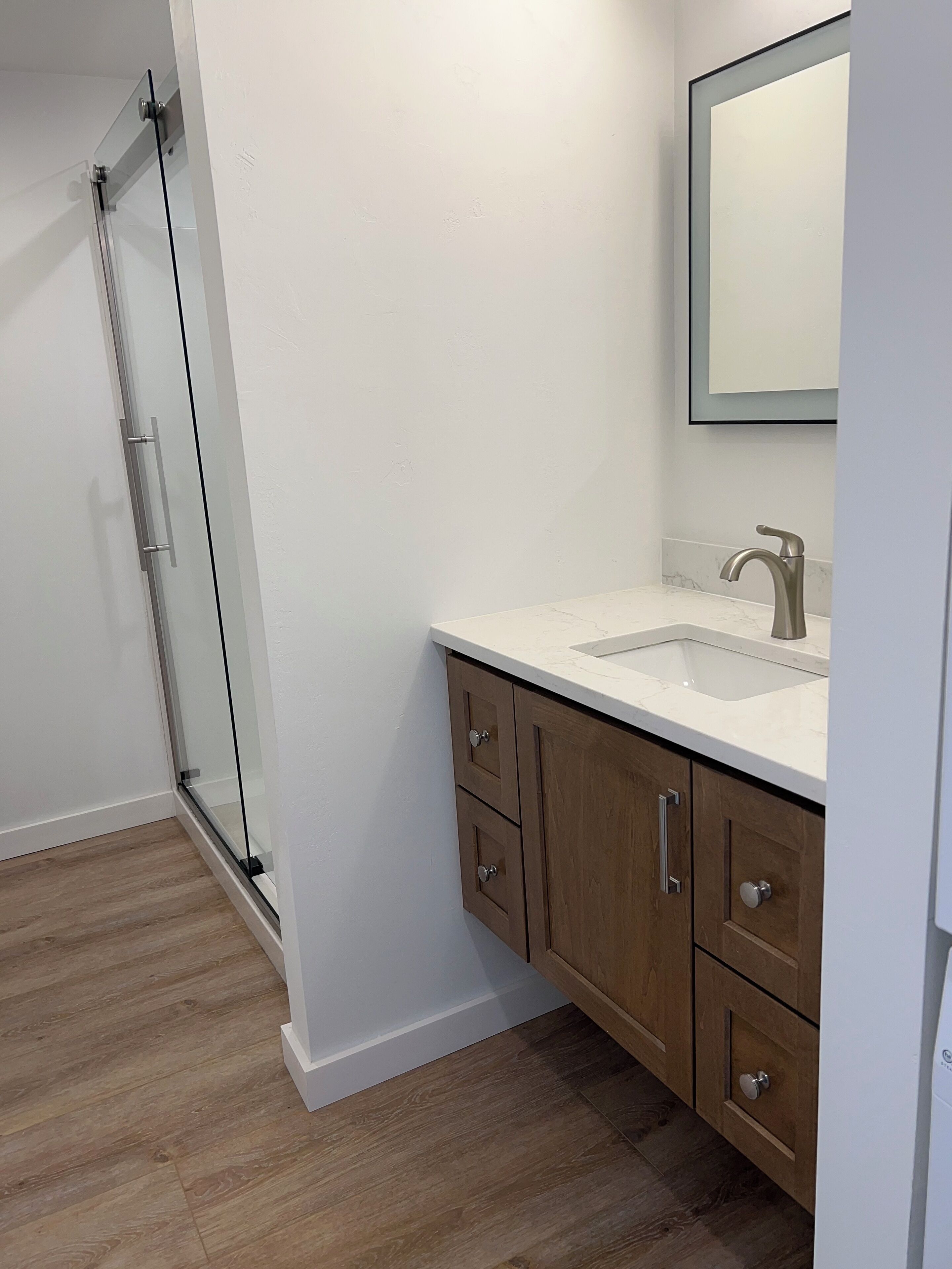 A bathroom with a glass shower enclosure, a sink with a marble countertop, wooden cabinets, and a mirror above the sink.