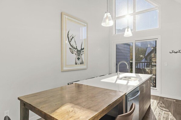 A modern kitchen with a wooden table, white countertop, and pendant lights.