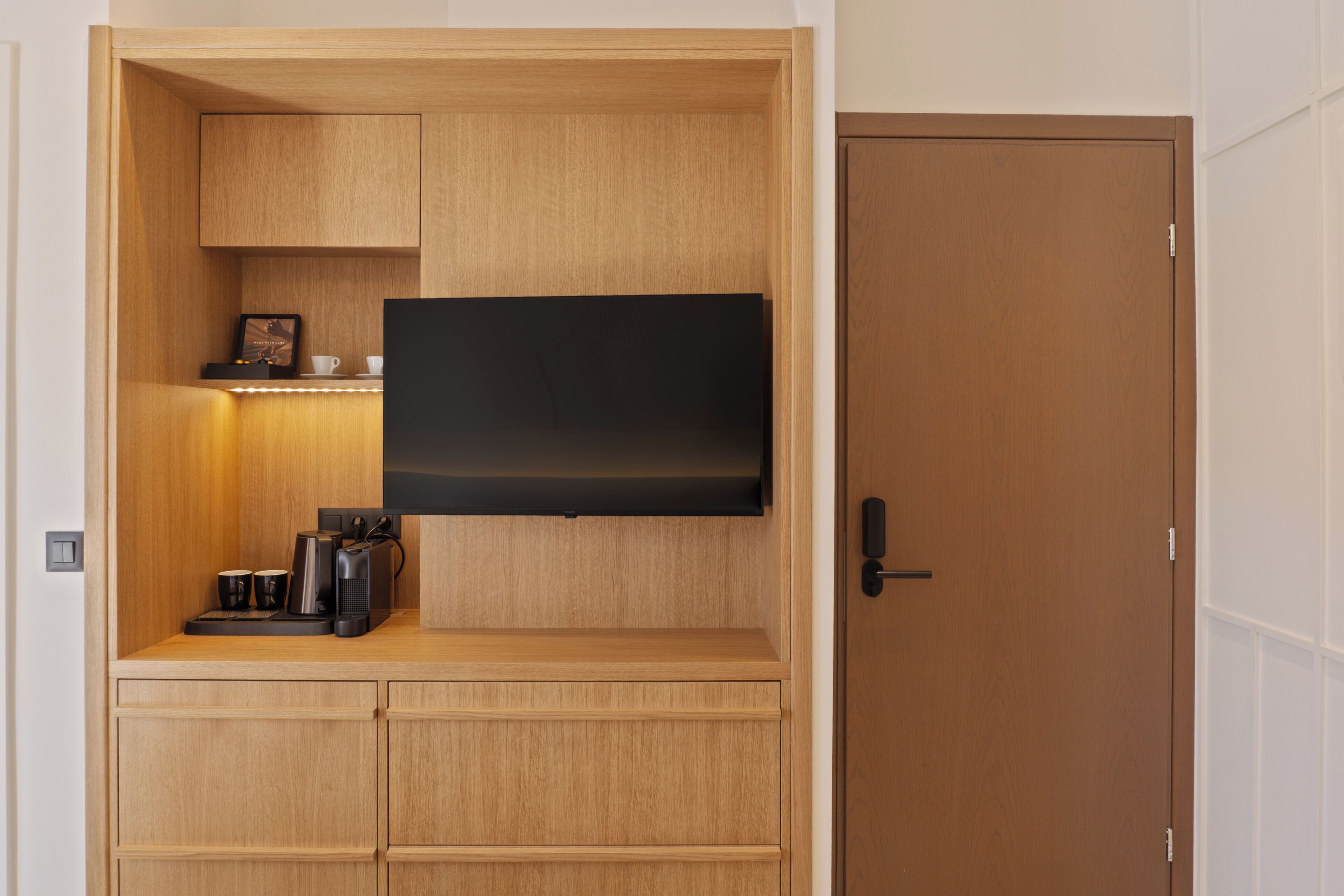 Una habitación compacta con una televisión de pantalla plana montada en un mueble de madera, una cafetera y una pequeña estantería con un marco de fotos.