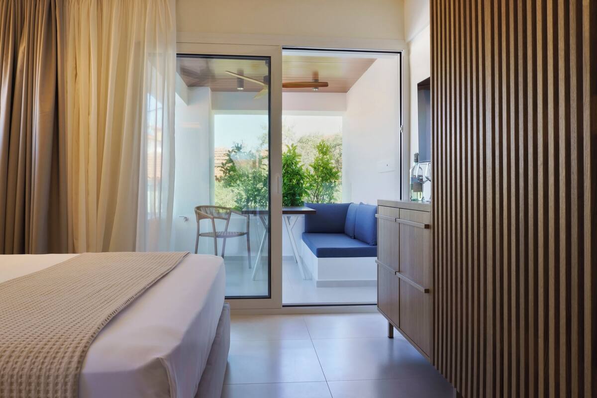 Habitación de hotel con cama, puerta corrediza de vidrio a un balcón, zona de estar con sofá y un mueble de madera.