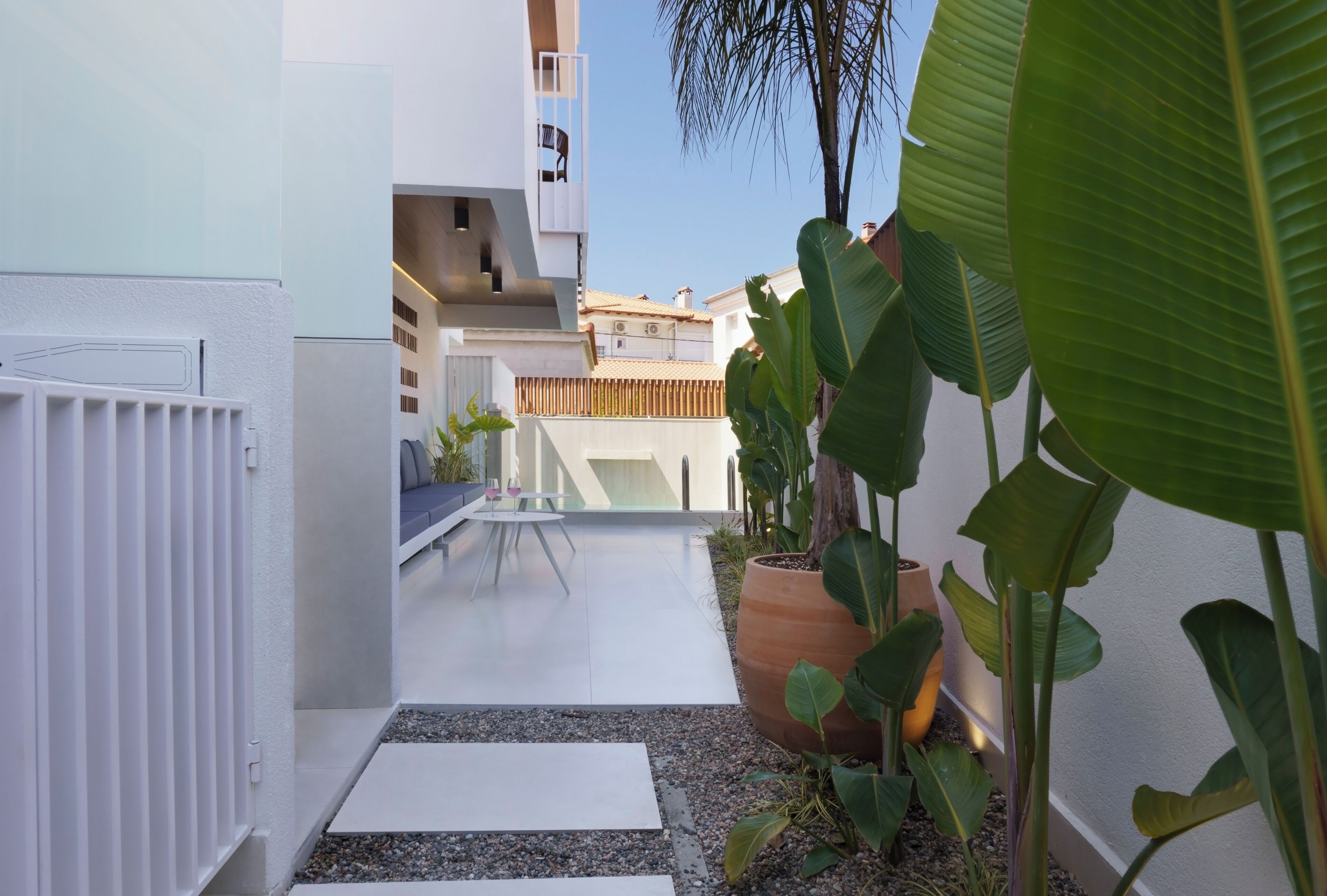 A narrow outdoor corridor with a white railing, a small table with chairs, and large potted plants.