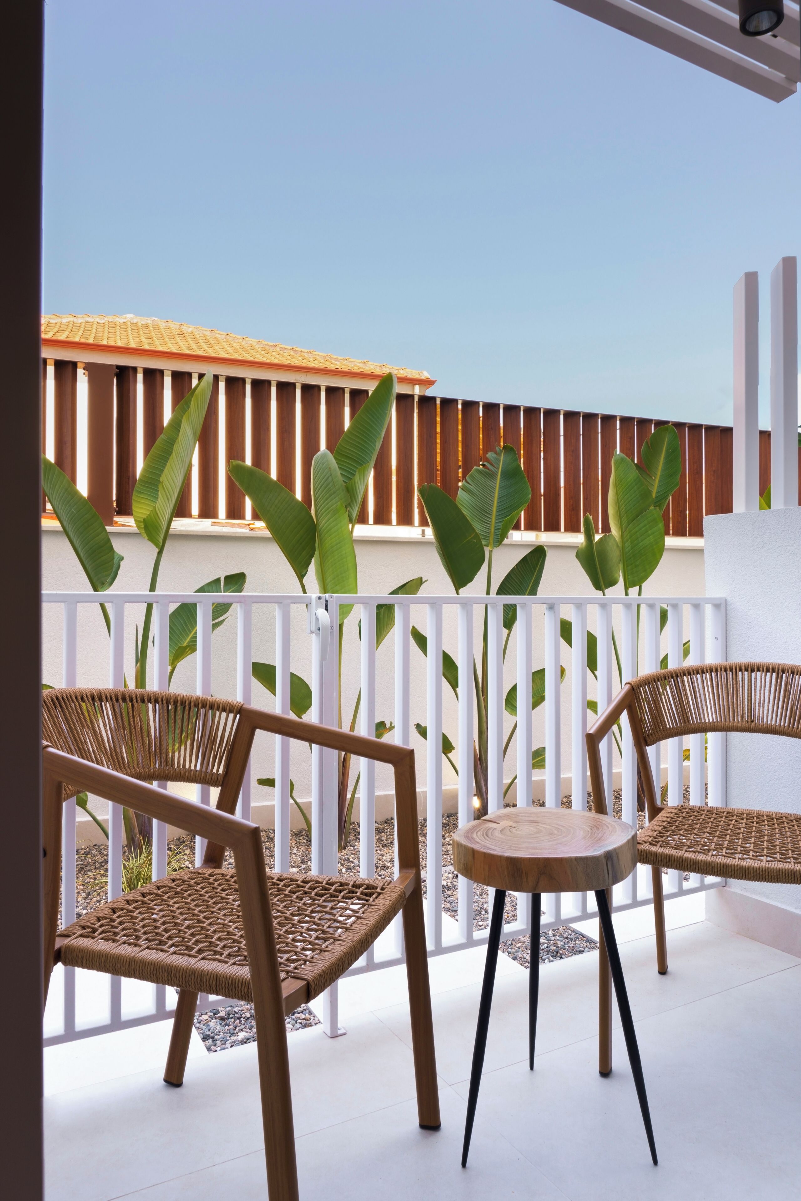 Een balkon met rieten stoelen, een ronde tafel, een witte balustrade en grote groene planten.