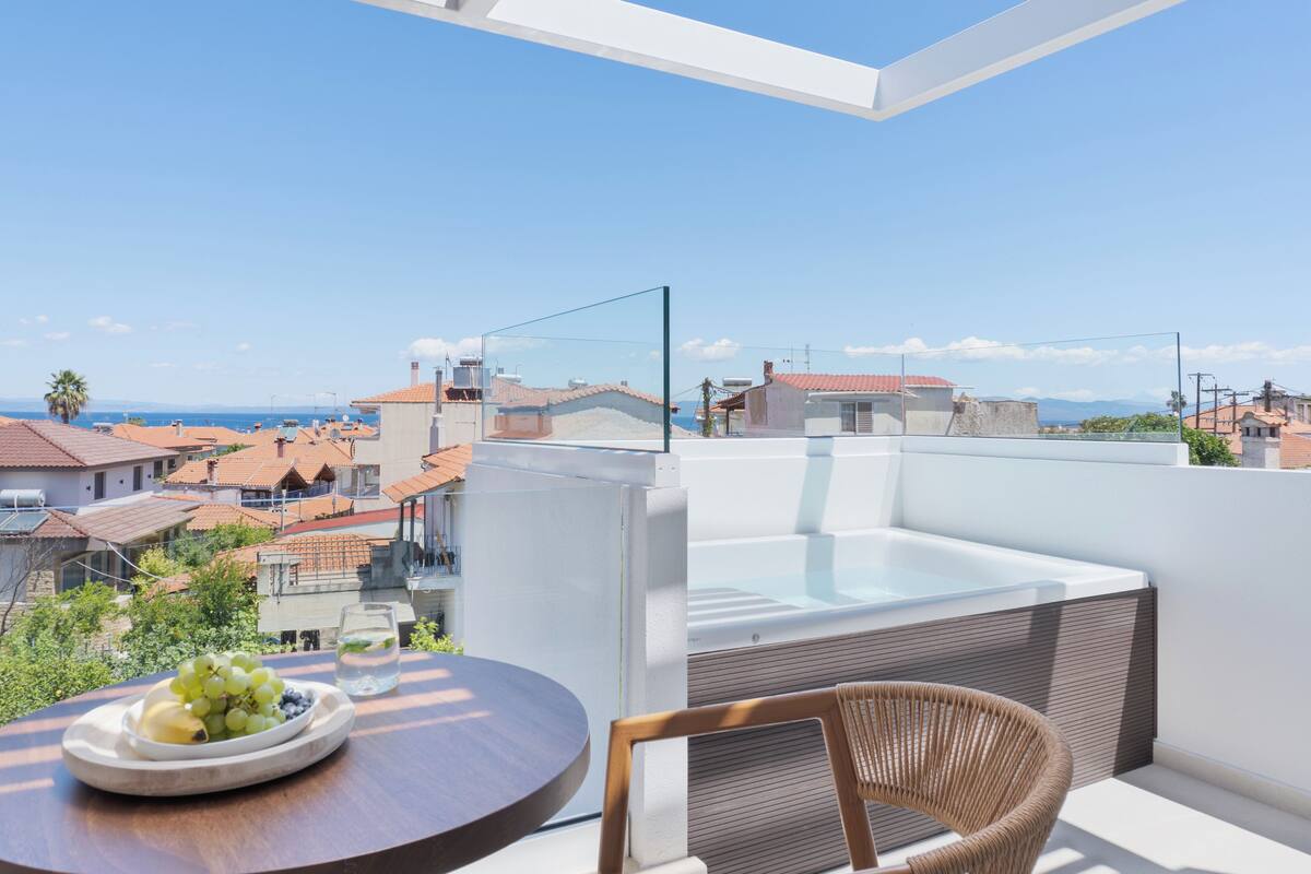 Un balcón con una barandilla de vidrio, un jacuzzi, mobiliario de mimbre y una mesa con fruta.