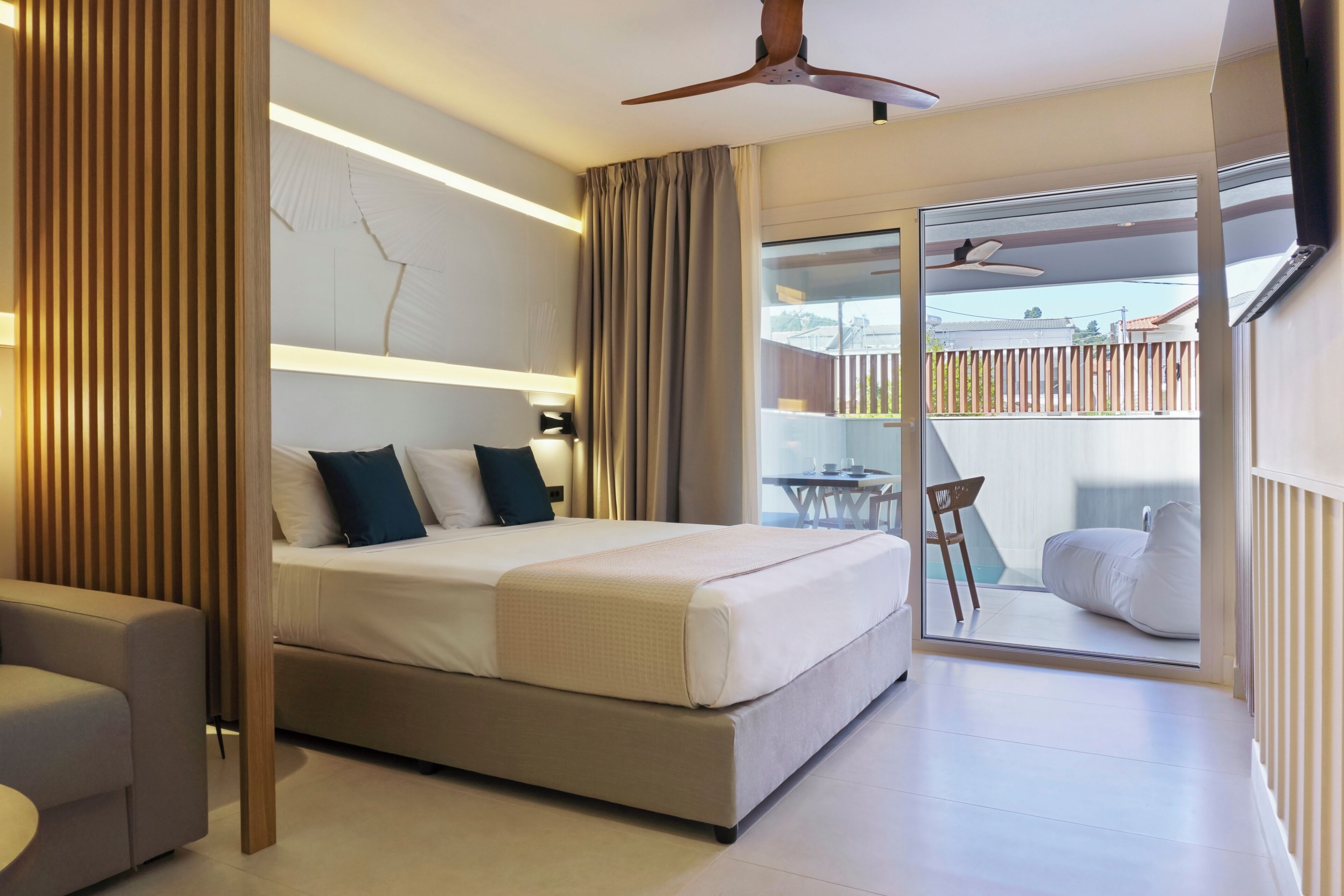 A modern hotel room with a large bed, a ceiling fan, a balcony with a table and chairs, and a view of the outside.
