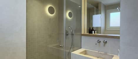 Un baño moderno con ducha envuelta en vidrio, un espejo grande y un tocador con lavamanos y espacio para toallas.
