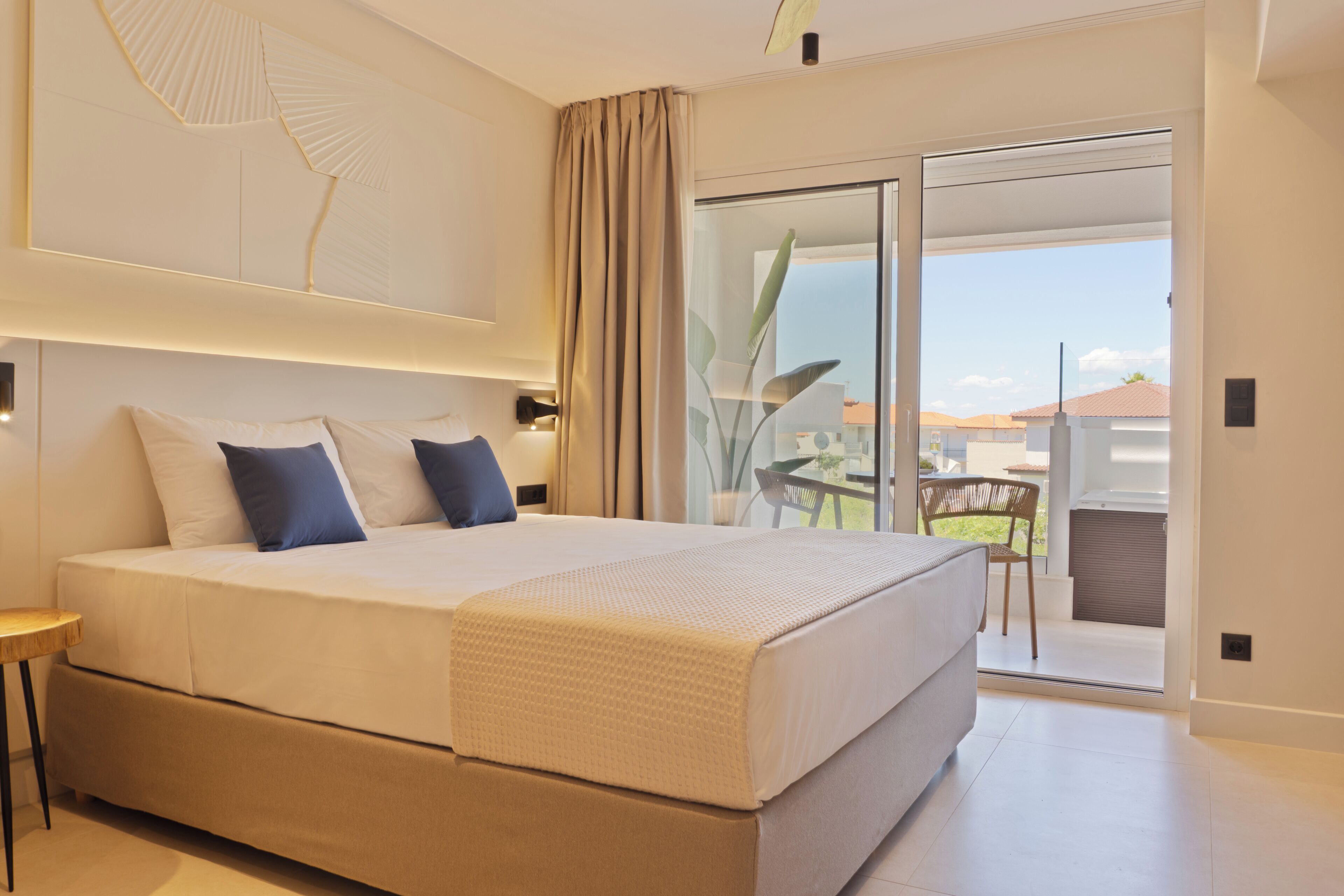 Una habitación de hotel moderna con una cama grande, una mesita auxiliar pequeña, una puerta corrediza de vidrio a un balcón y un elemento decorativo en la pared.