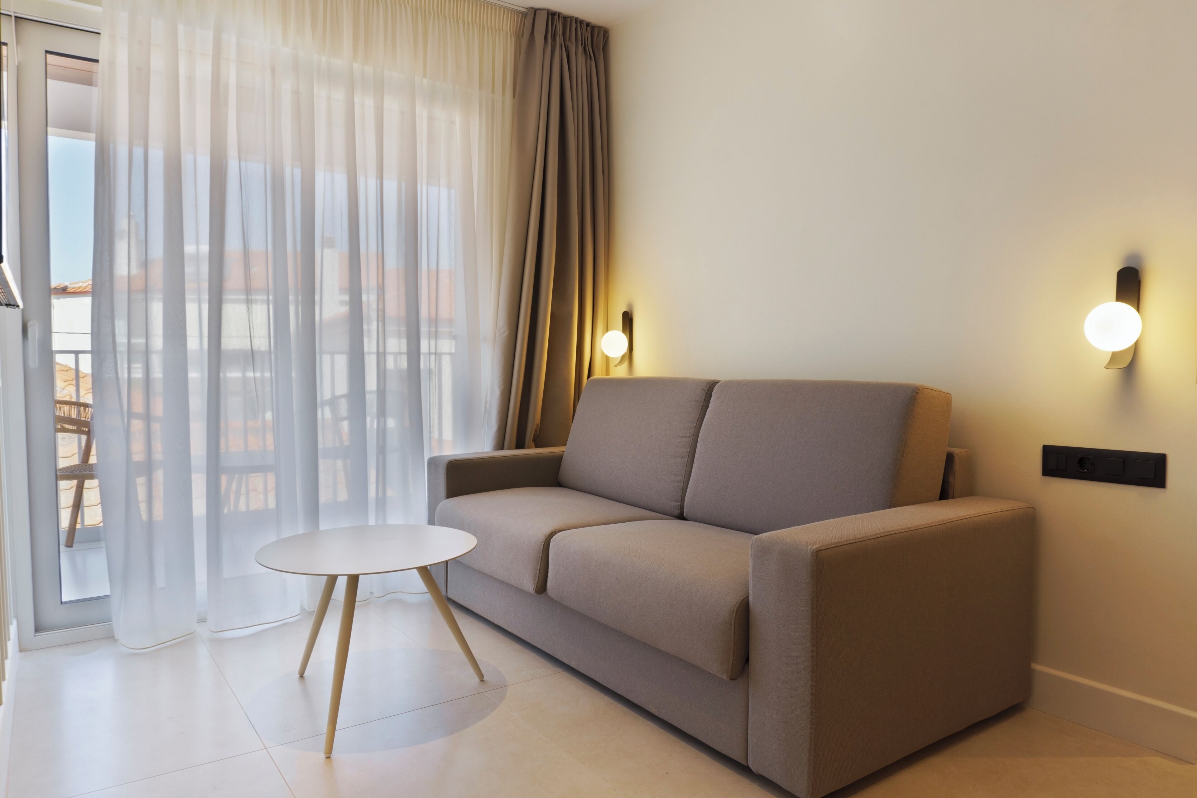 A modern living room with a sofa, a small round table, and a sliding glass door with sheer curtains.