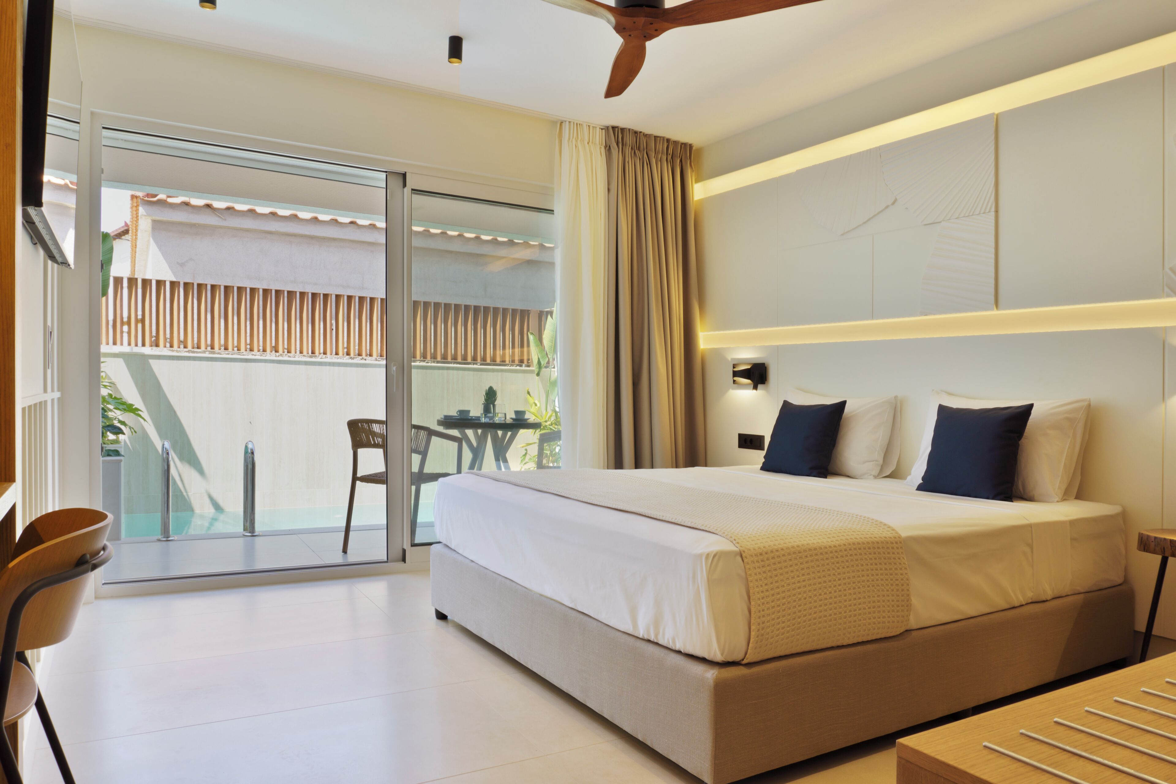 A modern hotel room with a large bed, a ceiling fan, a sliding glass door to a balcony, and a small table with chairs.