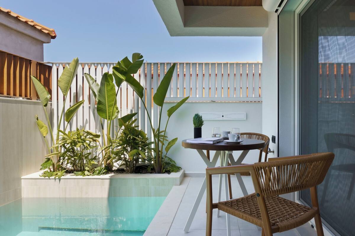 Zona junto a la piscina con una mesa y sillas, rodeada de exuberante vegetación y una valla blanca.