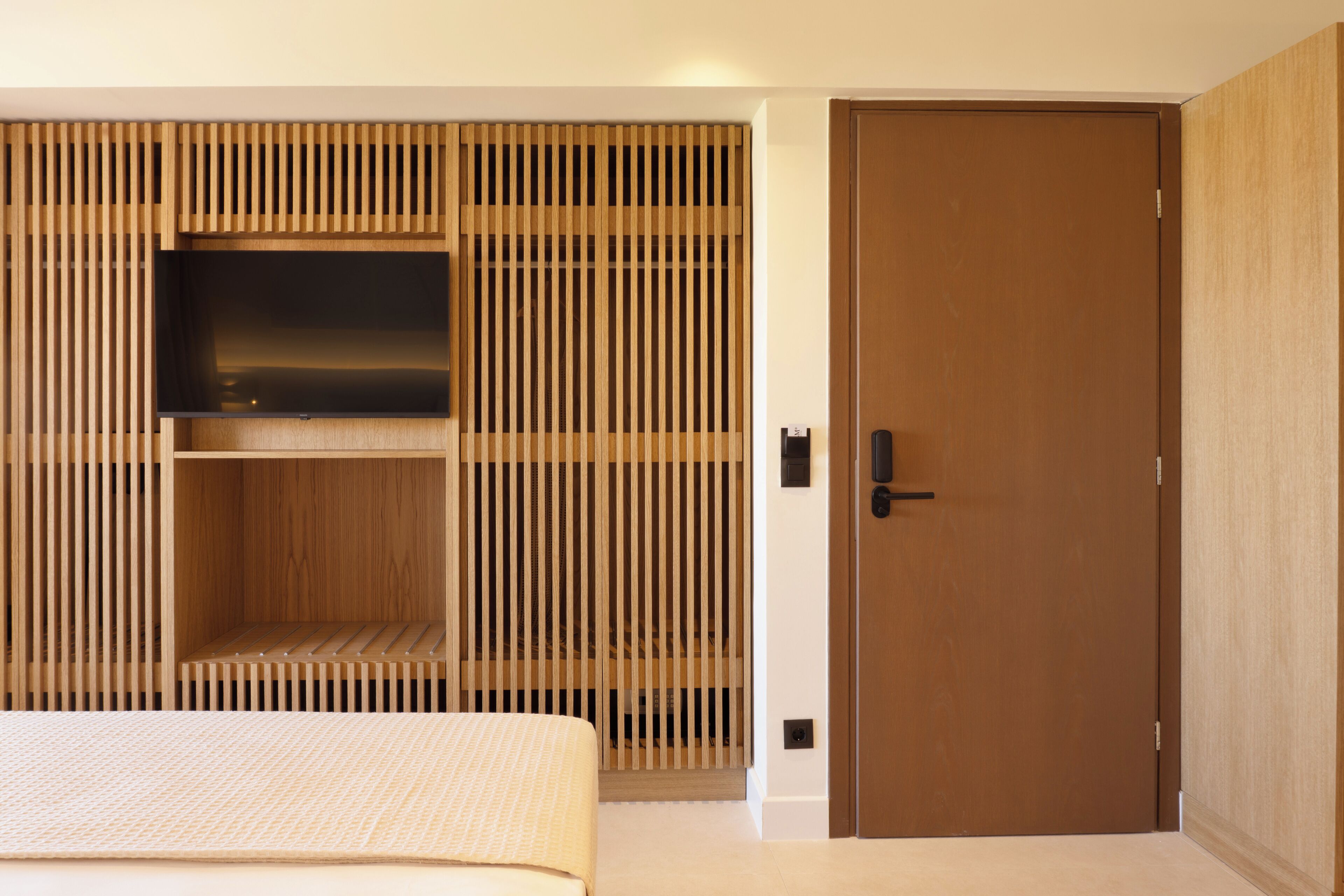 A modern bedroom with a bed, a TV mounted on a wooden slat wall, and a wooden door.