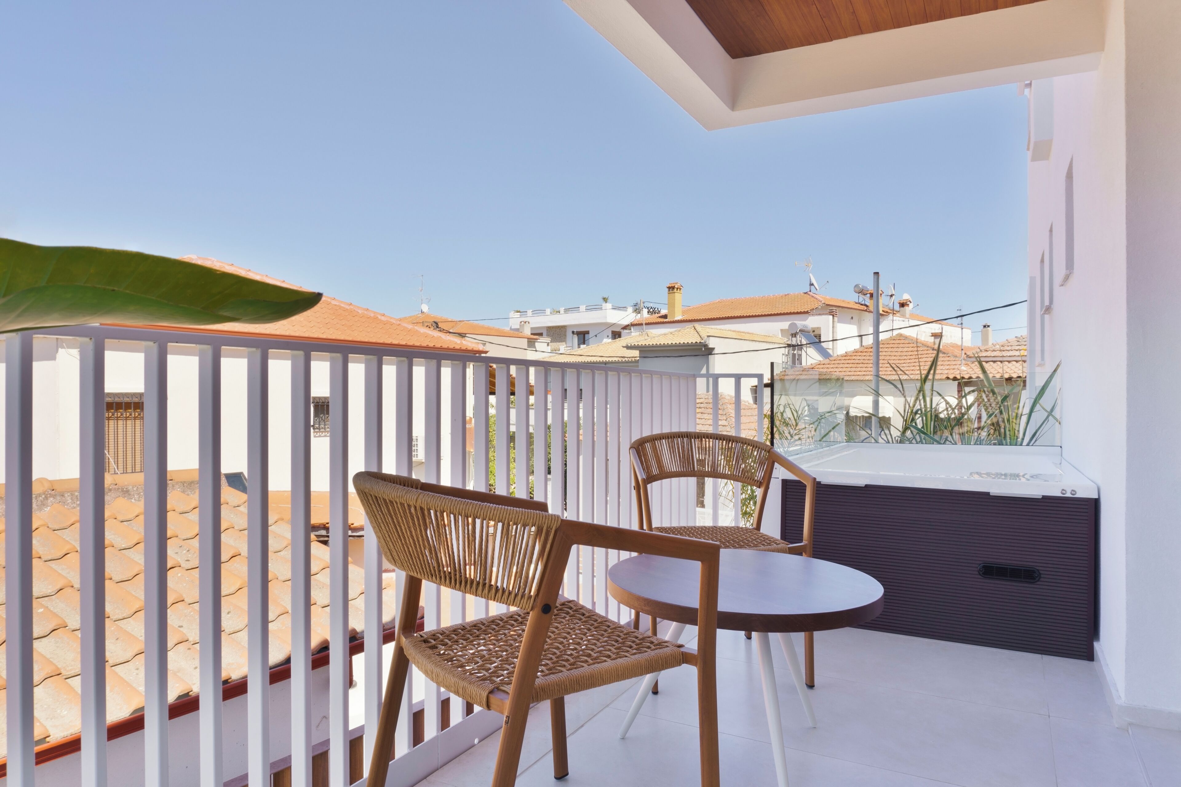 A balcony with two wicker chairs and a small round table, overlooking a tiled patio and neighboring buildings.