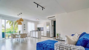 A modern living room with a blue sofa, a dining area with chairs, a kitchen with white cabinets, and a large window with a view of greenery. - 2Bdrm Apt. Front Juan Dolio Beach Access - Pool+BBQ (Juan Dolio)