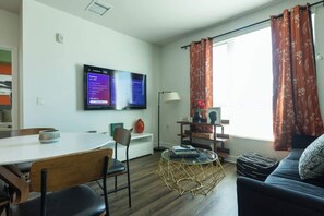 A modern living room with a flat-screen TV mounted on the wall, a dining table with chairs, a sofa, and a small coffee table. - Charming, Peaceful Apartment Retreat in Griffith (Glendale)