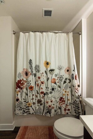 A bathroom with a shower curtain featuring a floral pattern, a toilet, and a small rug on the floor. - Charming, Peaceful Apartment Retreat in Griffith (Glendale)