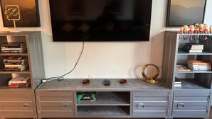 Een woonkamer met een flatscreen televisie aan de muur, een boekenkast vol boeken, een bijzettafeltje met decoratieve items, en een kleine kast met lades.