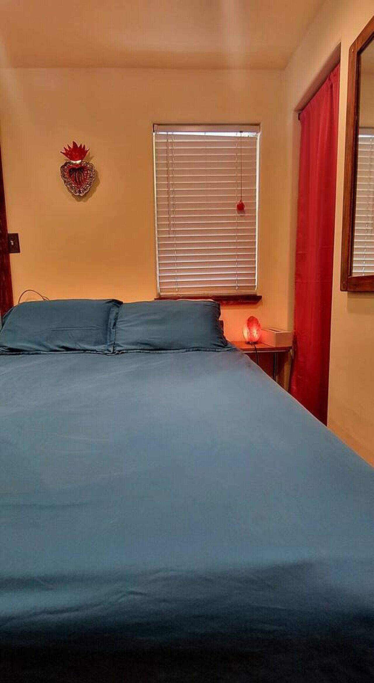 Une chambre à coucher avec un lit, une fenêtre avec des stores, un rideau rouge et un élément décoratif mural.