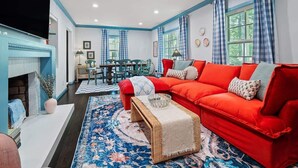 Un salon avec un canapé rouge, une table basse et un tapis bleu à motifs. On y trouve une cheminée, une table à manger avec des chaises et un téléviseur fixé au mur.