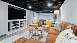Un salon moderne avec un canapé marron, des fauteuils jaunes et une table basse. Un téléviseur à écran plat est fixé au mur, et une petite table avec une lampe se trouve à proximité.