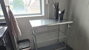 Una cocina con un carrito de acero inoxidable, un cubo de basura y una ventana que ofrece vistas a edificios.