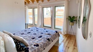 A bedroom with a sloped ceiling, a bed with patterned bedding, large windows, and a potted plant. - VIVA - Rooftop Terrace View Flat (Porto)