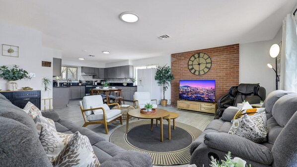 Ein modernes Wohnzimmer mit einem grauen Sofa, einem Couchtisch und einem Fernseher. Im Hintergrund ist eine Küche mit grauen Schränken und einer Ziegelwand zu sehen.