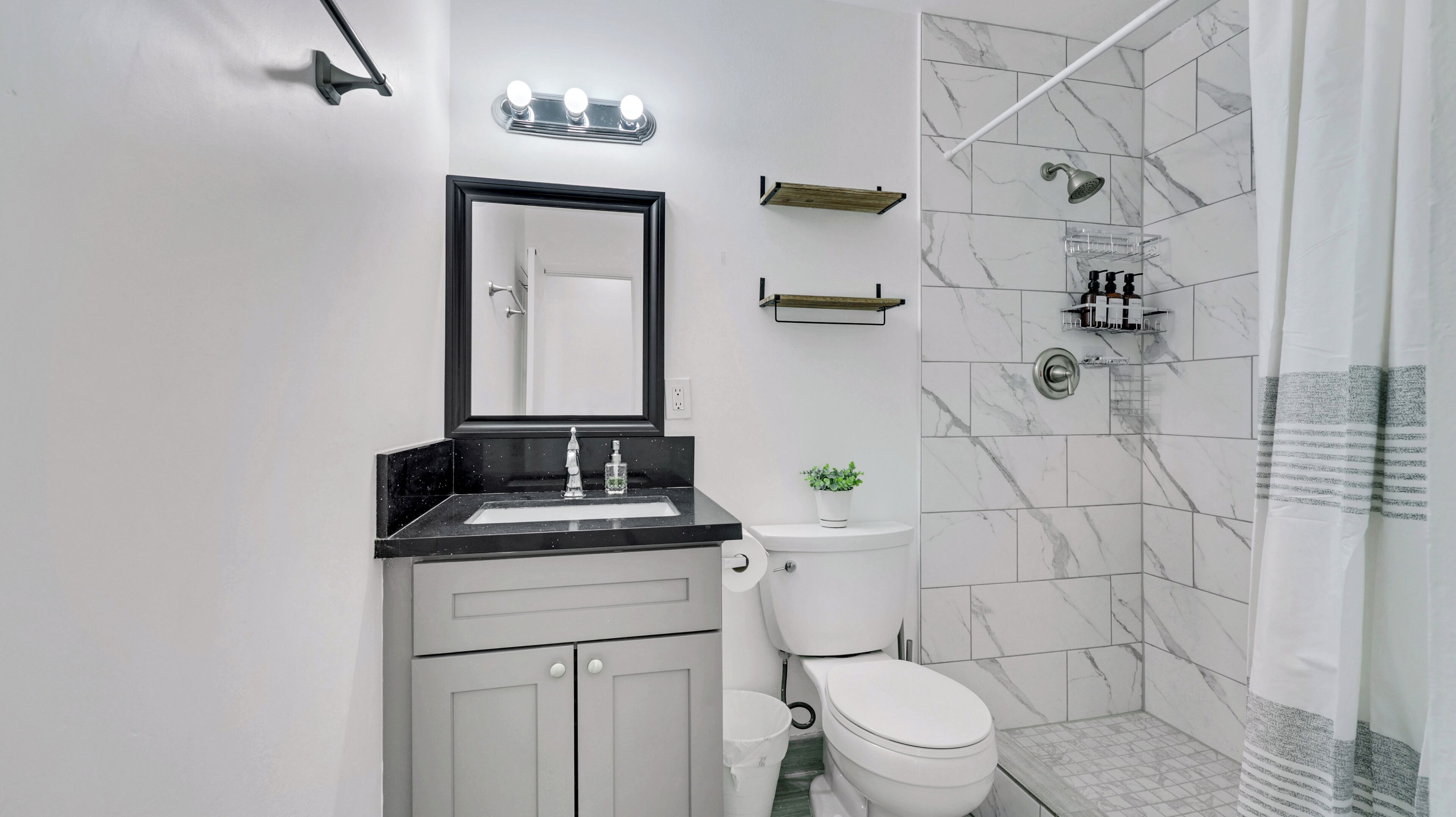 Une salle de bain moderne avec un lavabo sur un plan de toilette noir, des toilettes blanches, une douche équipée d’un pommeau de douche à main et un espace de douche avec des carreaux de marbre.