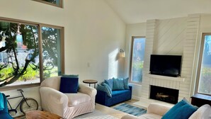 A modern living room with a fireplace, a television, and a large window with a view of greenery. - Perfect 2-bedroom beach condo in Oxnard (Oxnard)