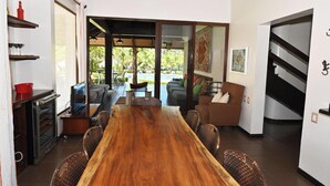 A spacious dining area with a long wooden table, wicker chairs, and a view of a pool and greenery outside. - Villas Estival Playa Prieta (Tempate)