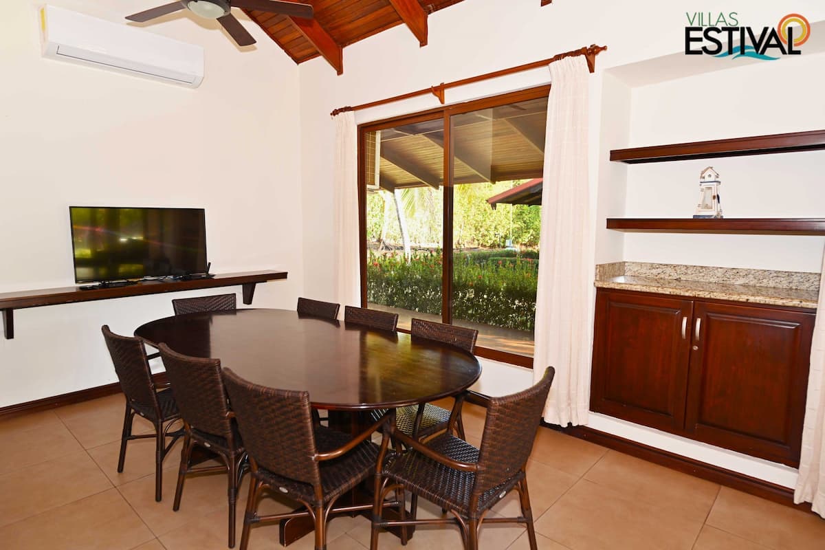 Un comedor con una mesa redonda y sillas, un televisor y vistas al exterior con vegetación.