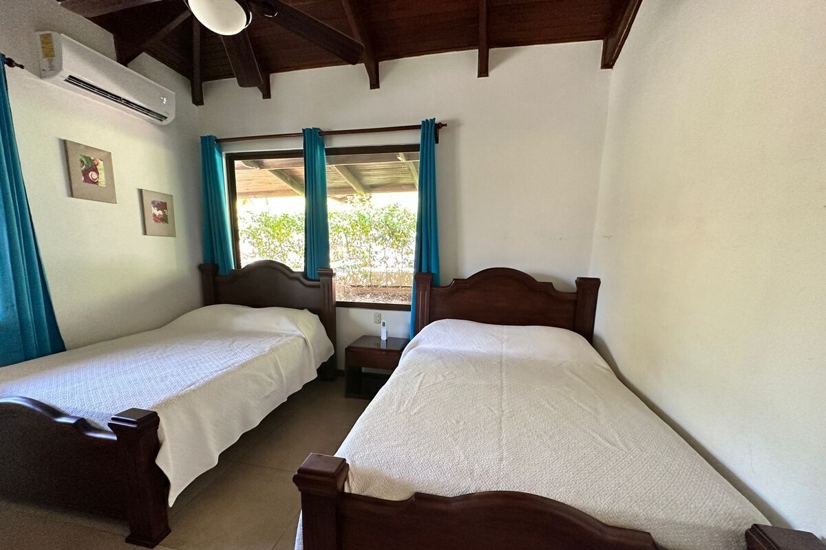 A room with two wooden beds, a window with curtains, and an air conditioning unit on the wall.