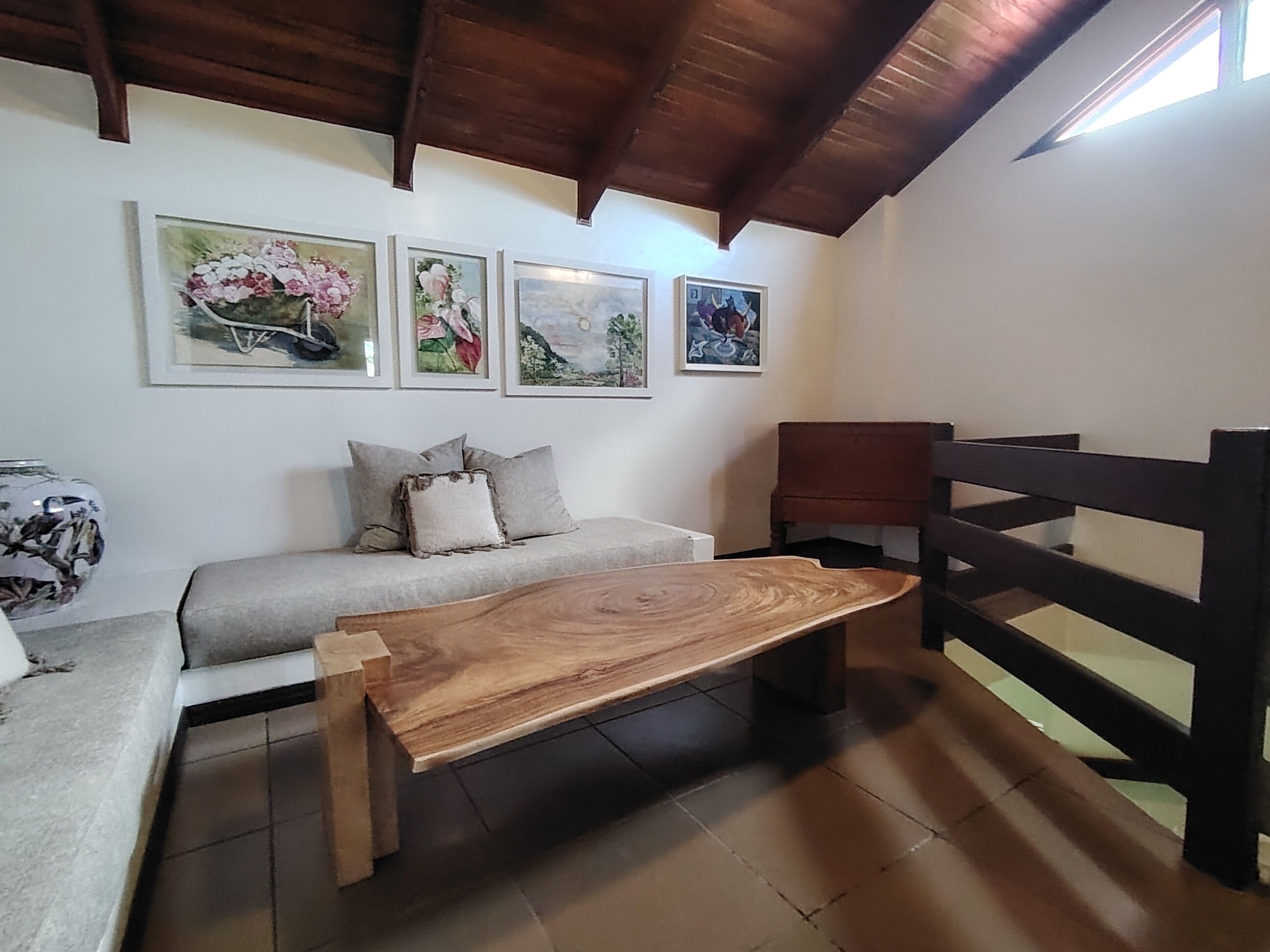 A cozy room with a wooden bench, a sofa with cushions, and a wooden chair. There are three framed artworks on the wall.