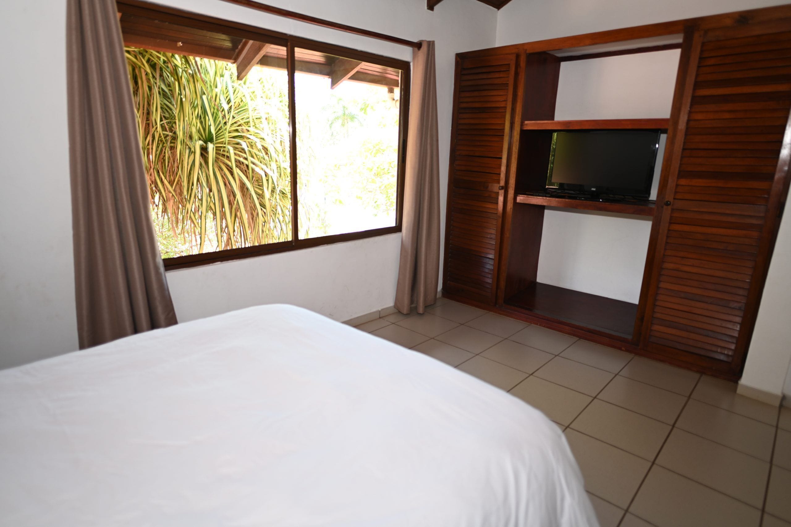 Quarto com cama, televisão sobre uma prateleira de madeira e janela com cortinas.