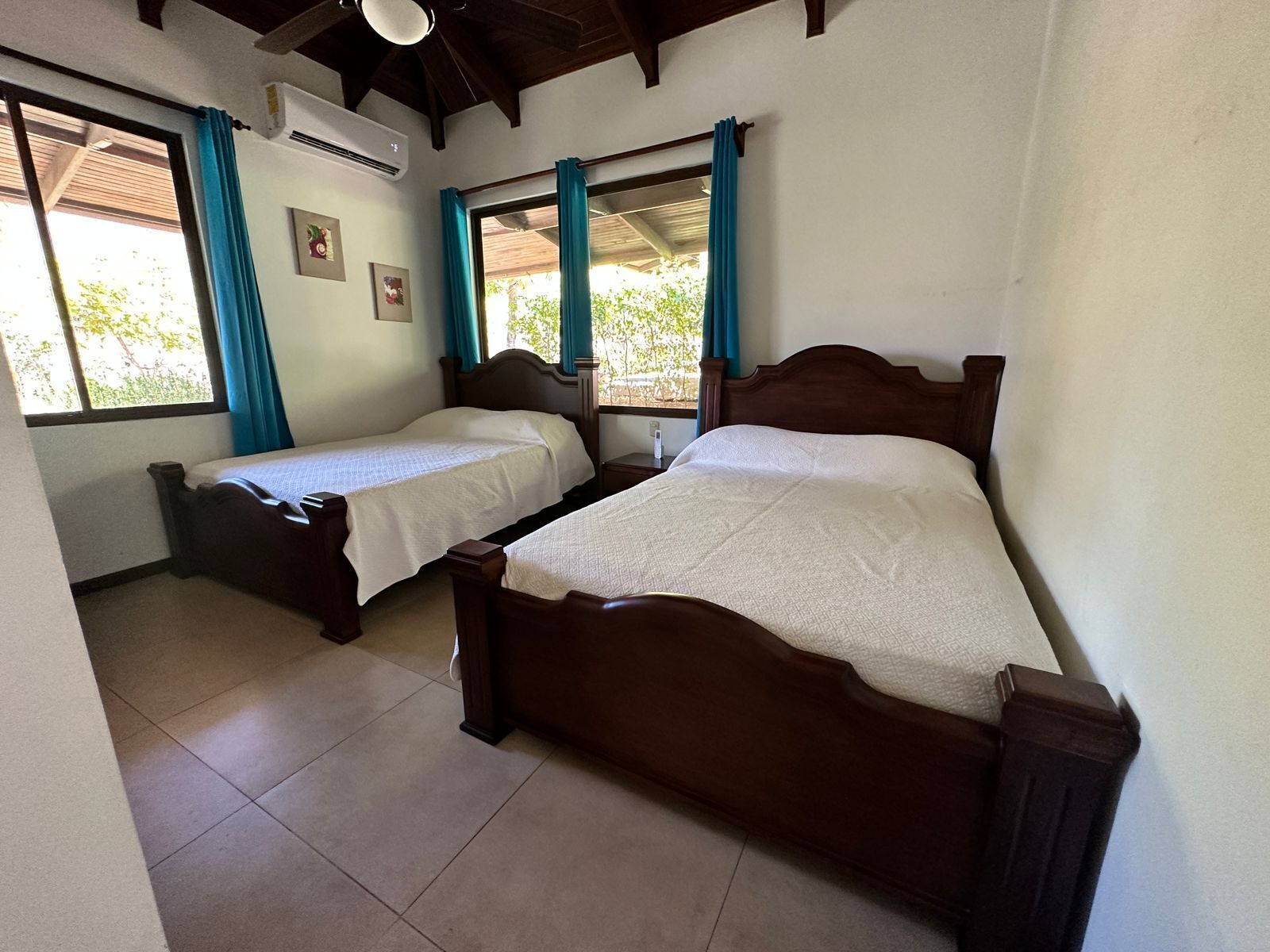 Une chambre avec deux lits en bois, une fenêtre avec des rideaux et un sol carrelé.