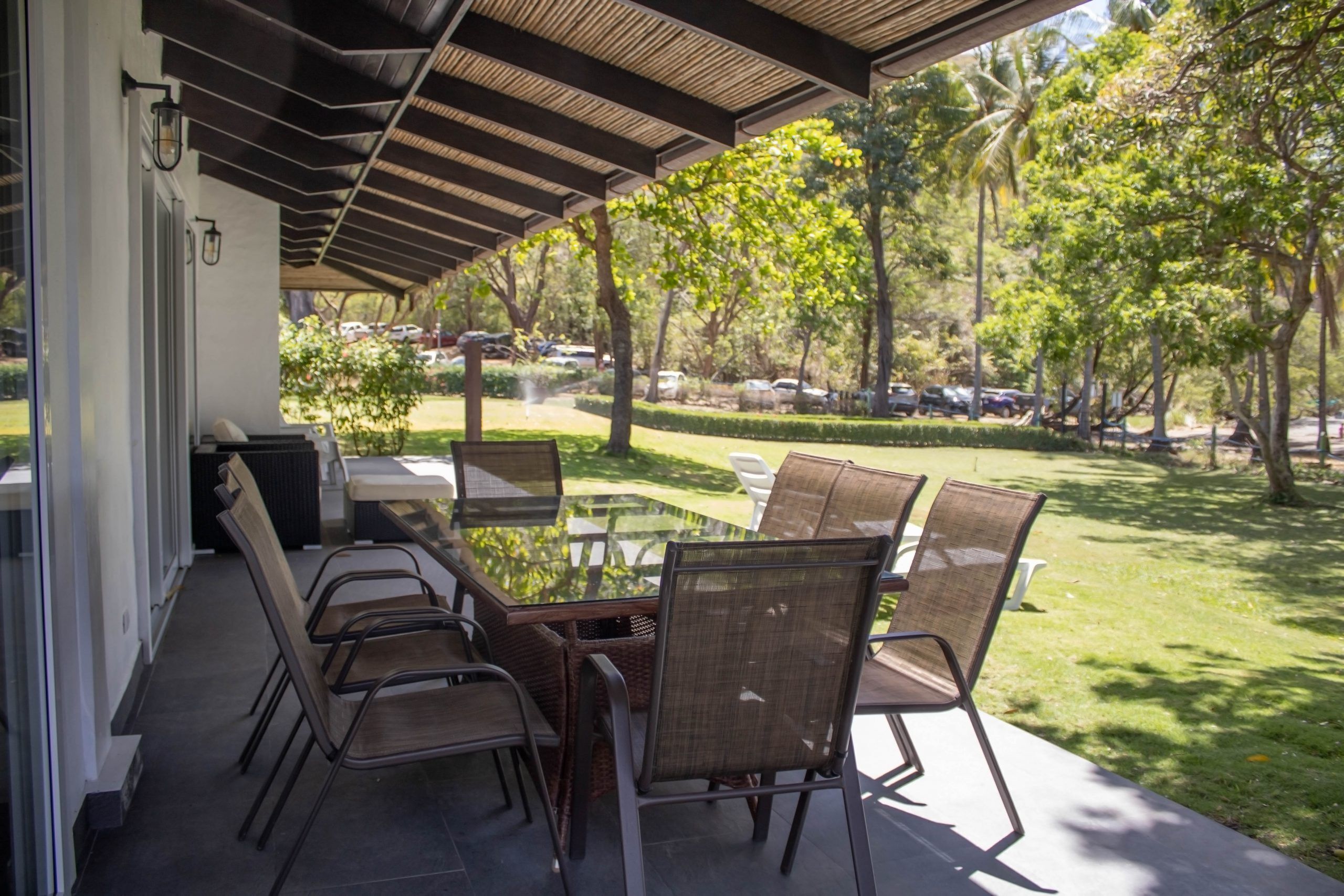 An outdoor patio with a glass table and wicker chairs, overlooking a well-maintained lawn and parked cars.