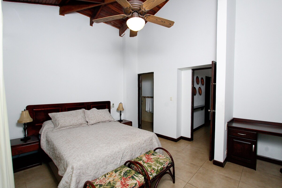 A bedroom with a wooden bed, a nightstand, a bench, a desk, and a ceiling fan.