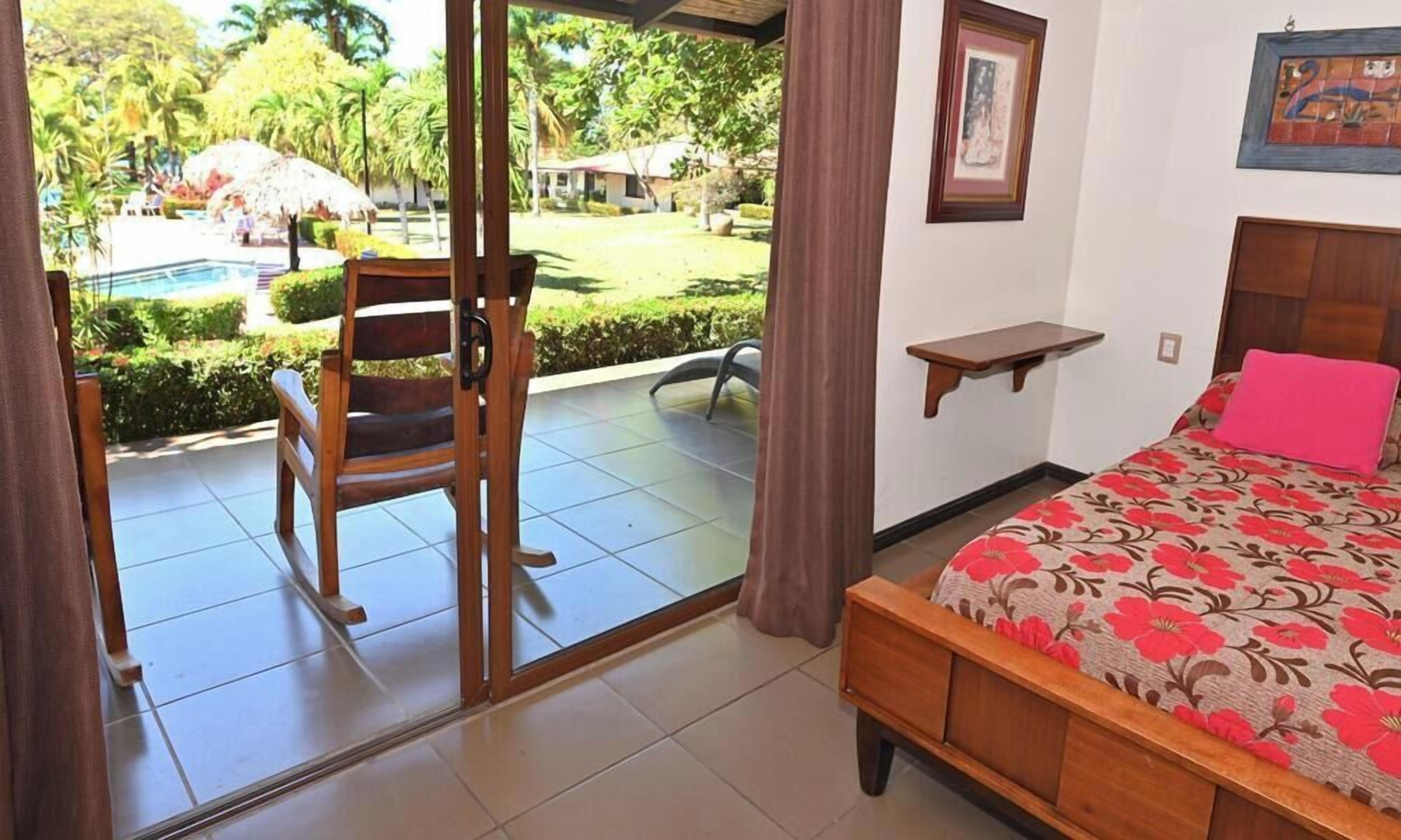 A hotel room with a bed, a small table, a chair, and a view of a pool and garden.