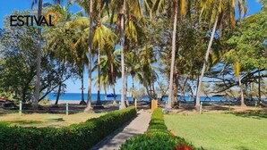 A pathway lined with palm trees and red flowers leading to a beach with umbrellas and a clear blue sky. - Villas Estival Playa Prieta (Tempate)