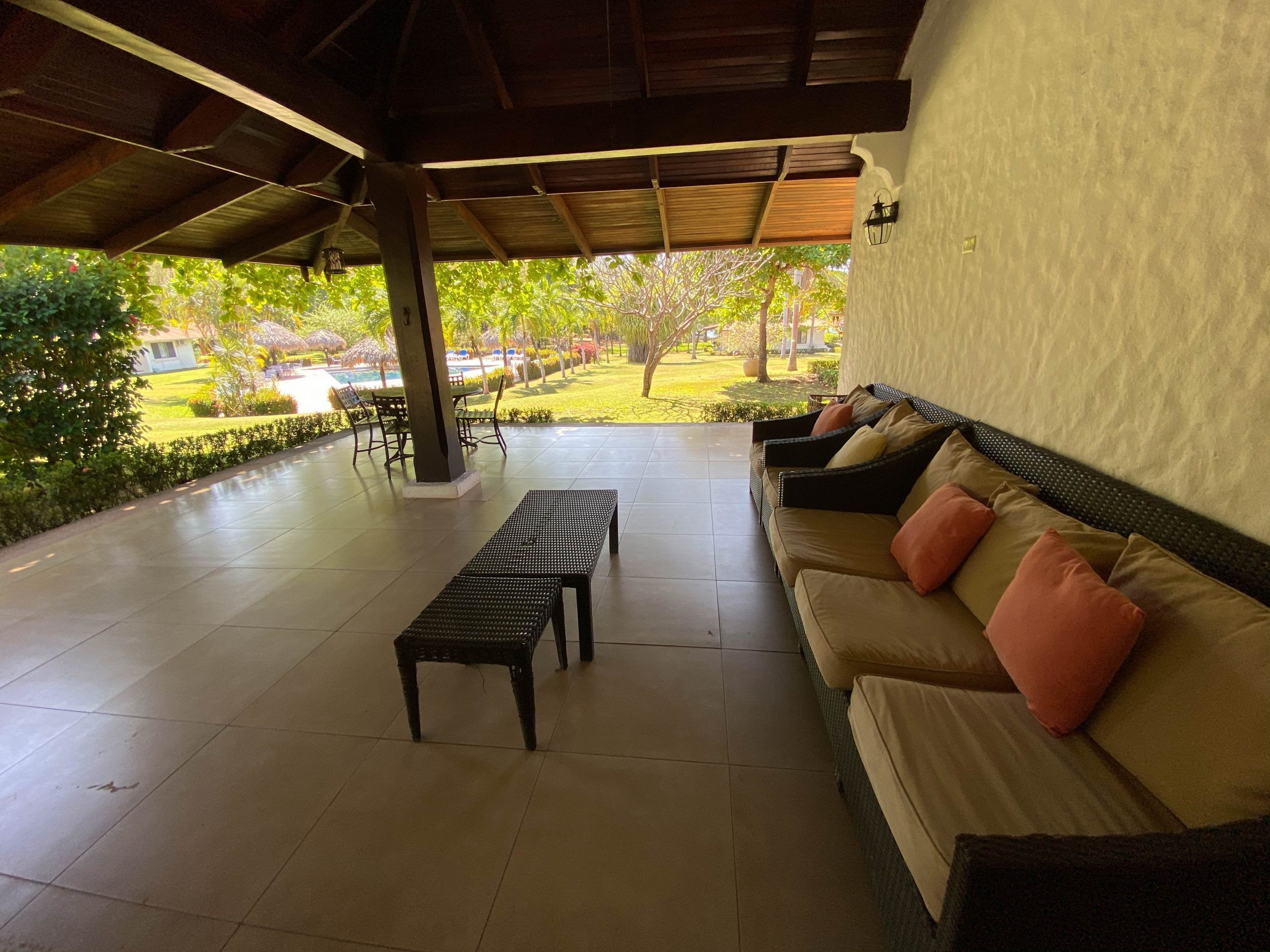 Un espacio de estar al aire libre cubierto con sofá y banco, suelo de baldosas y vistas a un jardín con piscina y árboles.