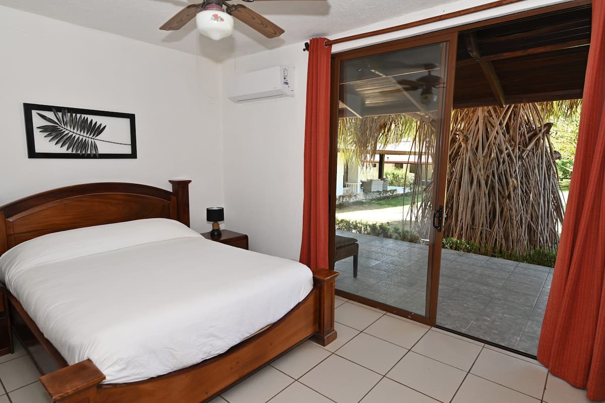 Un dormitorio con una cama de madera, una mesita de noche, un ventilador de techo y un ventanal corredero que da a un espacio exterior.