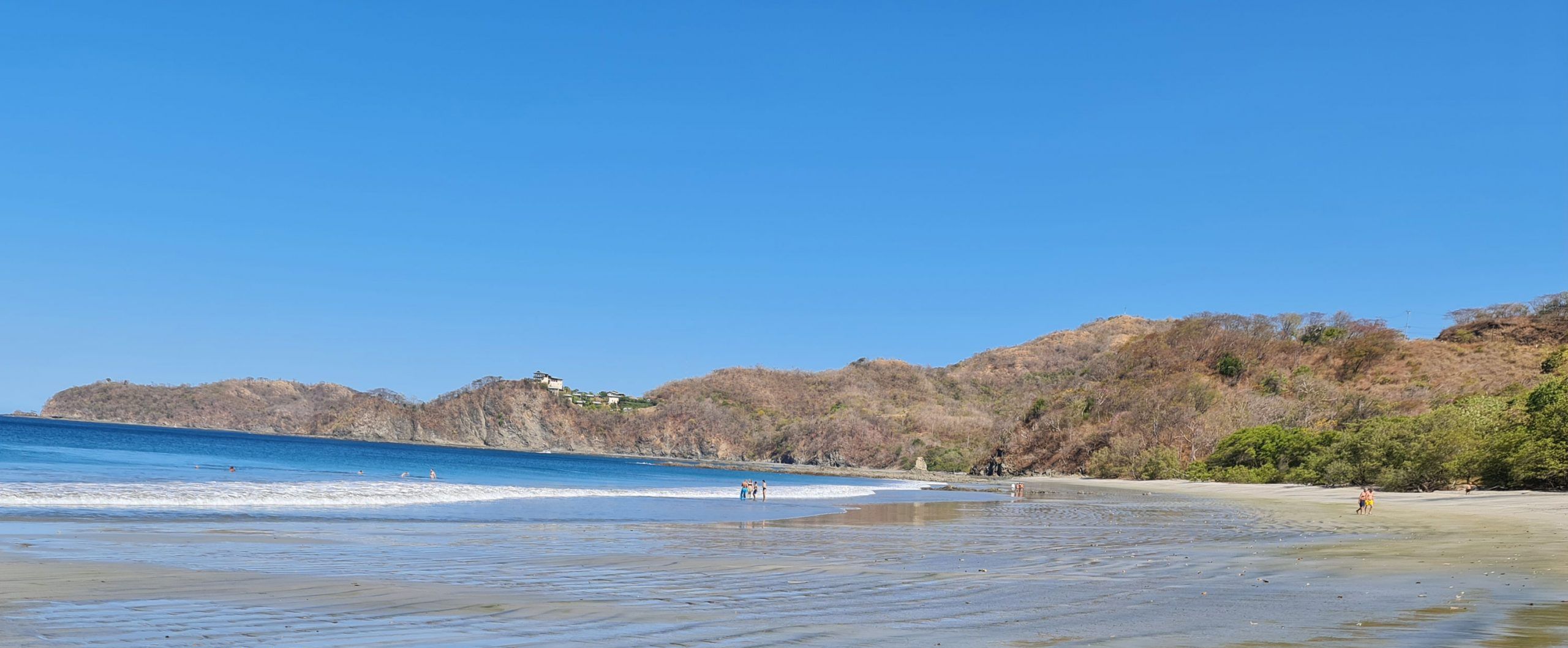 A beach with clear blue water, a sandy shore, and a hill with sparse vegetation in the background.
