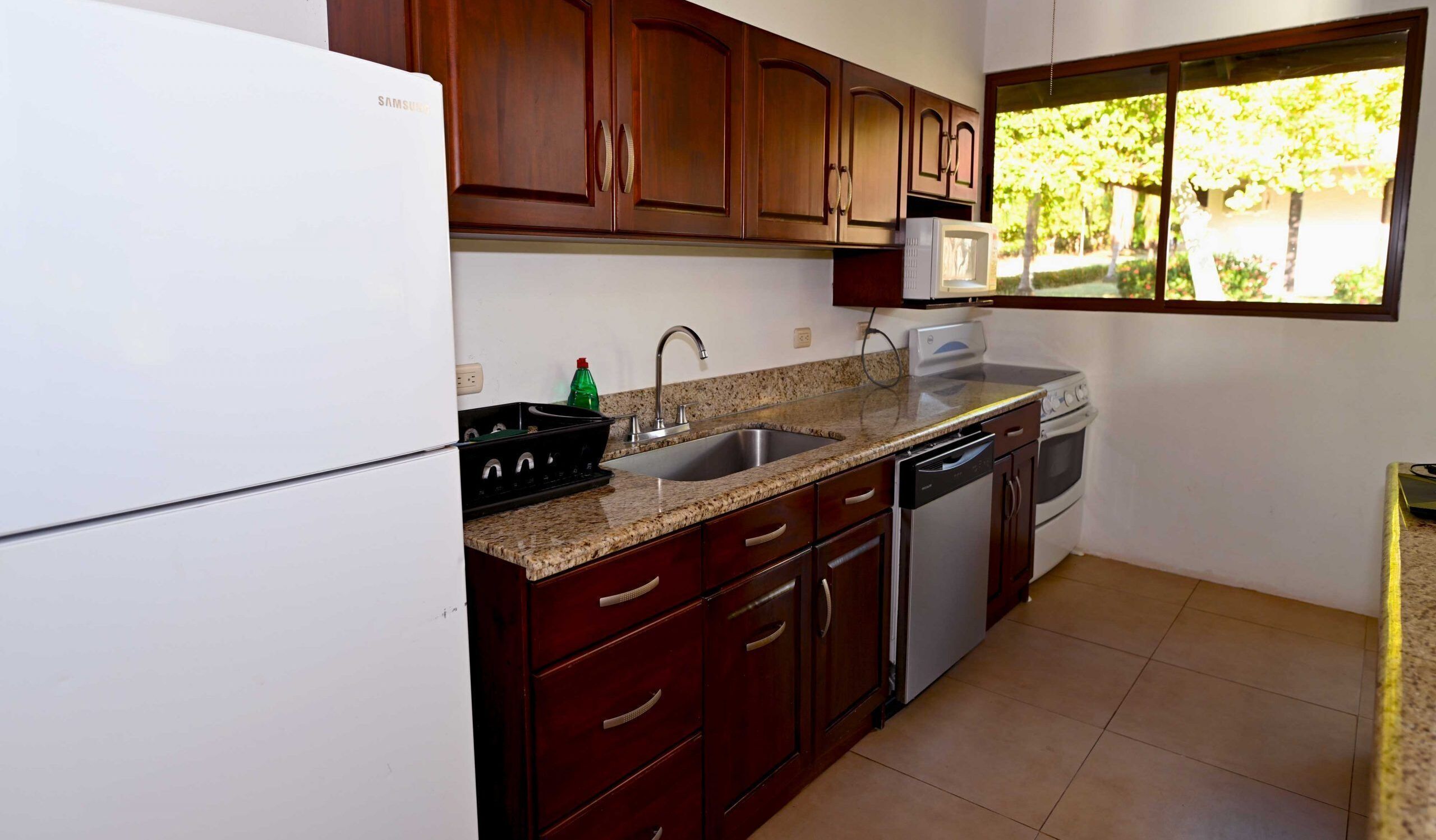 Una cocina con armarios de madera, un lavamanos de acero inoxidable, lavavajillas y microondas.