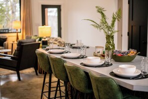Comedor con una larga mesa dispuesta para seis comensales, sillas tapizadas en verde, un jarrón con follaje verde y un bol de aperitivos.