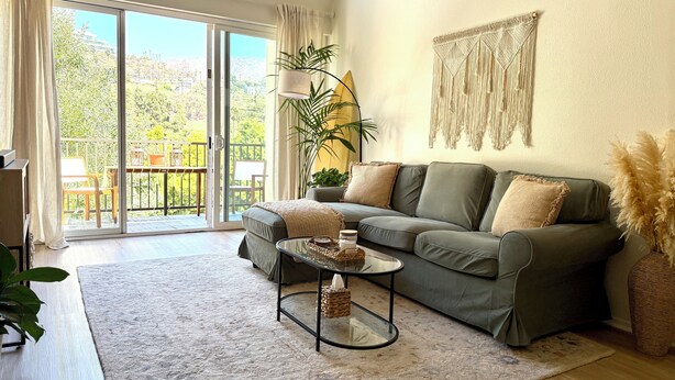 A living room with a grey sofa, a glass coffee table, a potted plant, and a view of a balcony with outdoor furniture. - Beachy Malibu Loft - Pool / Tennis / Gym (Malibu)