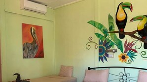 Una habitación con cama, dos mesitas de noche, ventilador de techo, aire acondicionado instalado en la pared y un mural con pájaros y plantas.