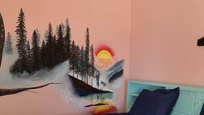 Un lit avec une couverture beige, une tête de lit turquoise et une fresque murale représentant une forêt et un soleil.