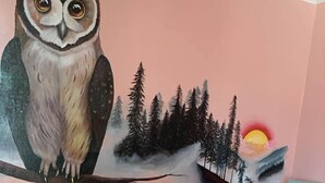 Un lit avec un matelas bleu et une couverture blanche, un mur rose orné d’une fresque représentant une chouette et une forêt, et un plancher en bois.